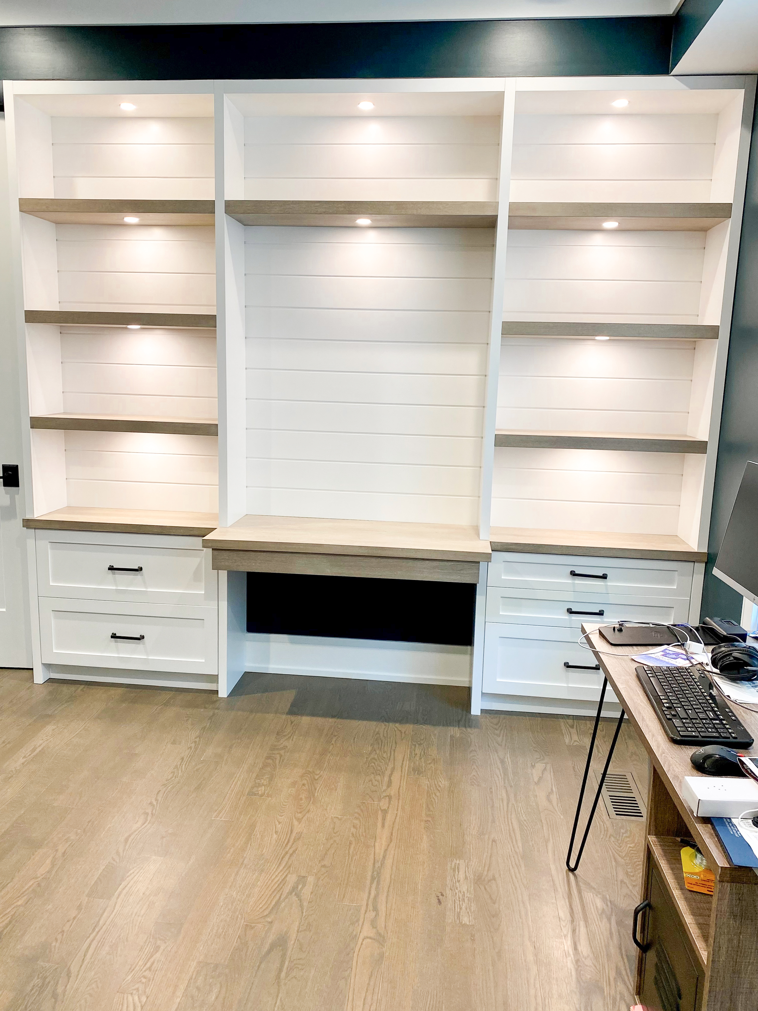 White painted home office built-in with shiplap back panel, three open shelf bays, LED lighting, and integrated desk surface