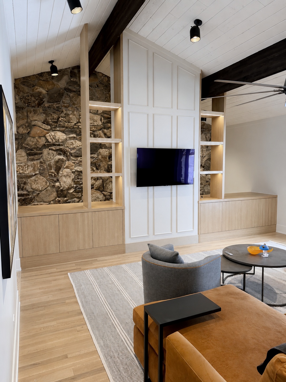 White oak media wall built-in with open shelving flanking a white panel wall, set against a stone accent wall in a vaulted living room