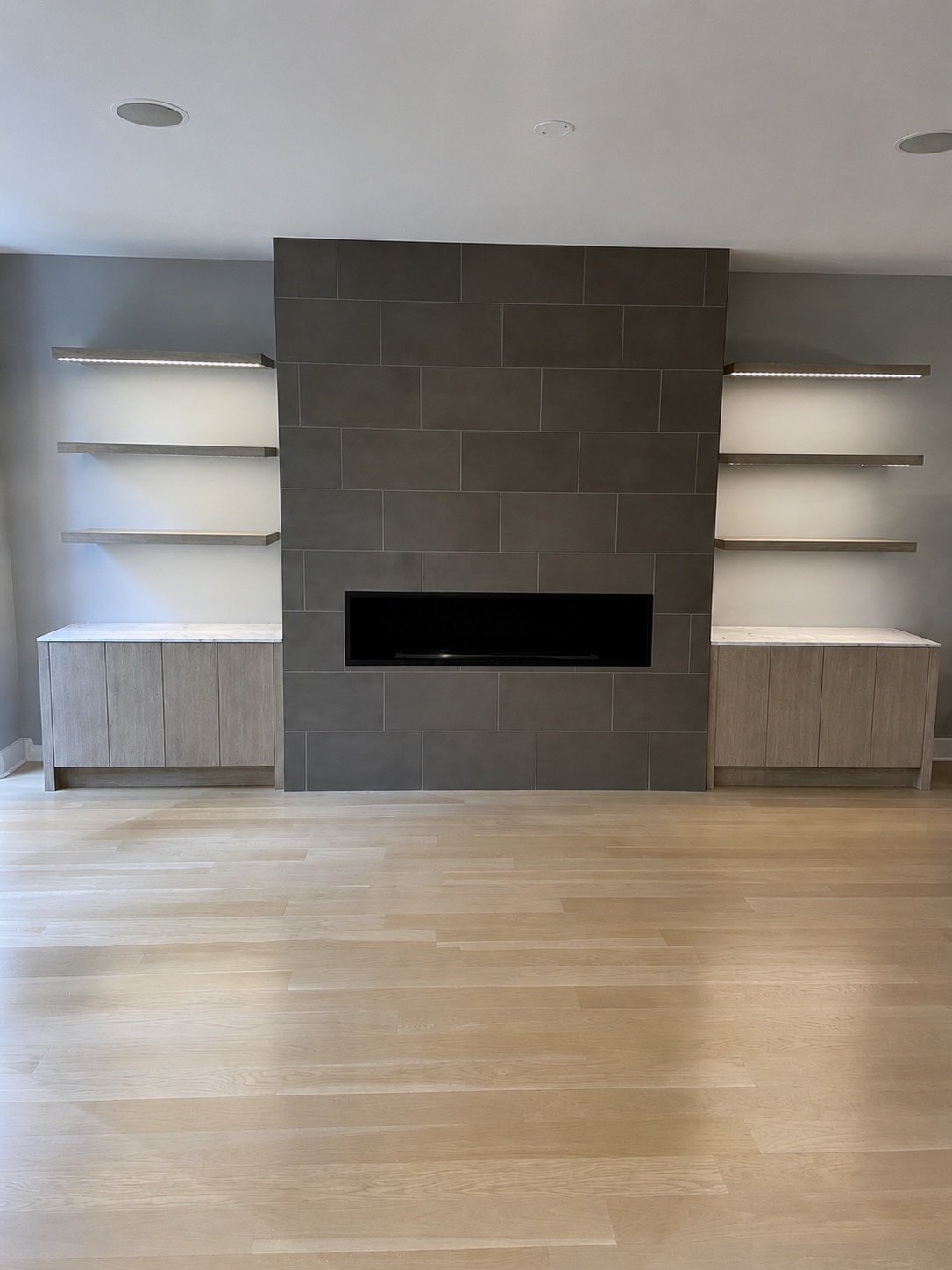 Modern gray tile fireplace wall with flanking white oak lower cabinets, marble countertops, and floating LED shelves