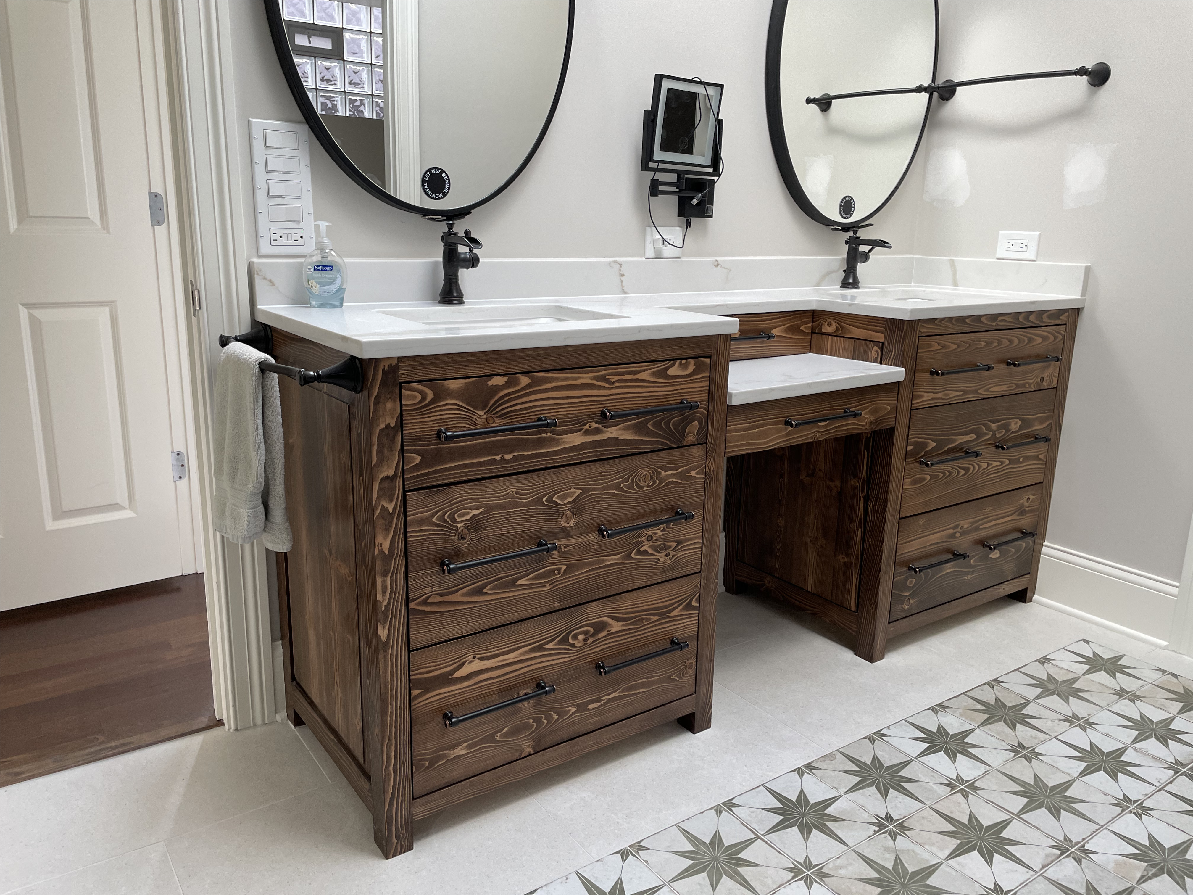Dark stained double vanity split by a center makeup area, with black fixtures and star mosaic tile floor