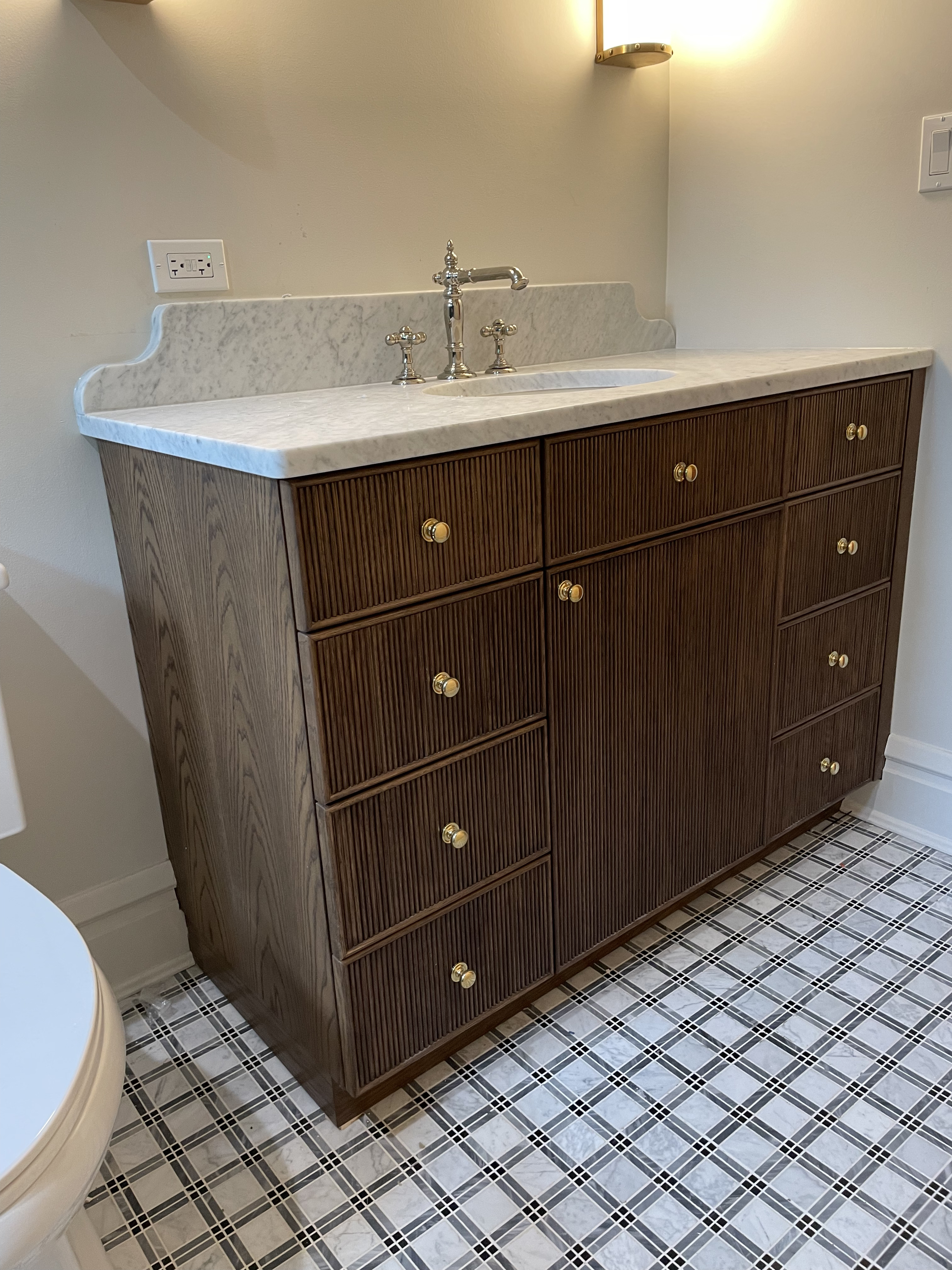 Dark wood fluted single-sink vanity with marble top, shaped backsplash, and bridge faucet on plaid tile floor