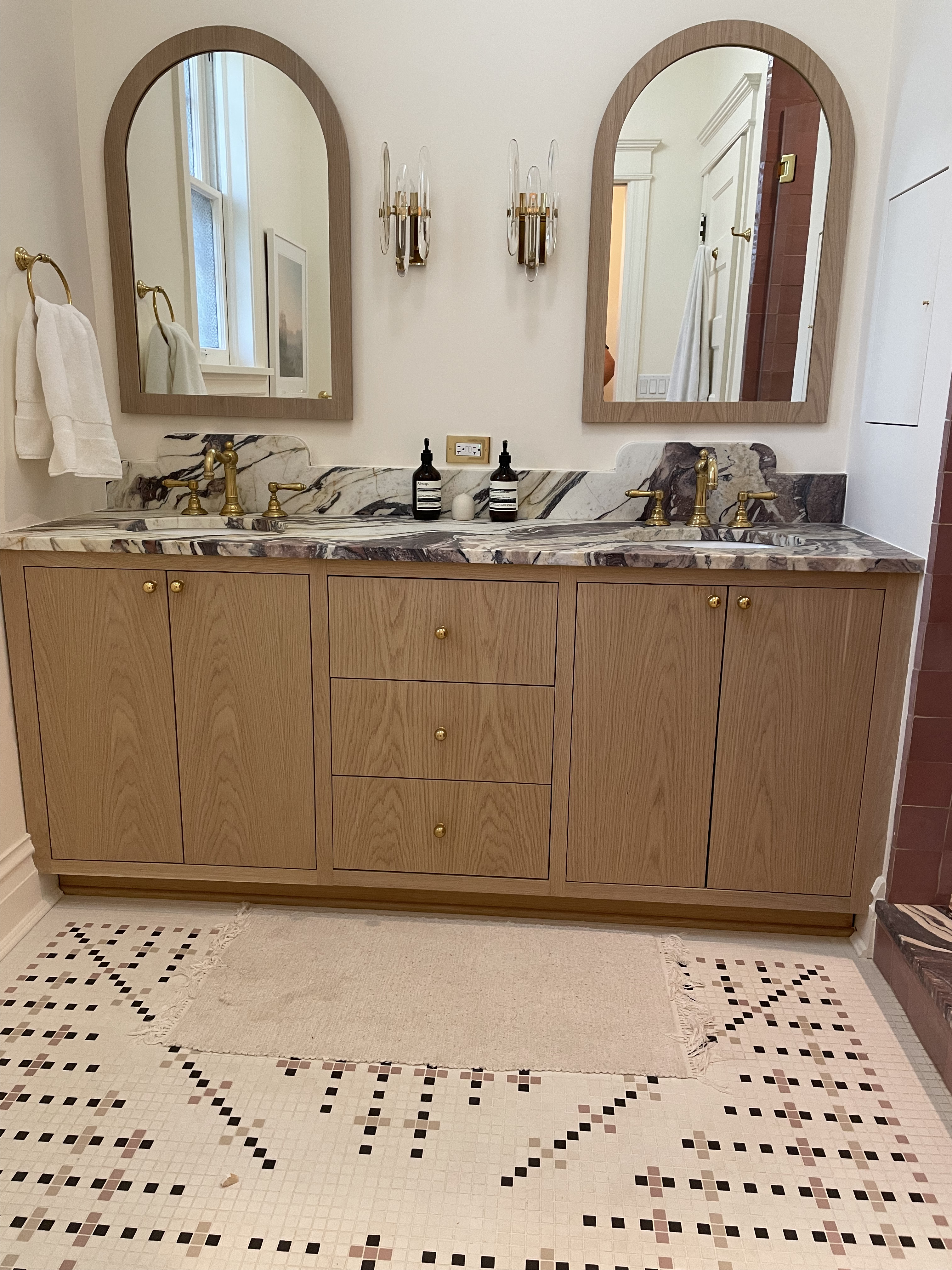 White oak double vanity with purple waterfall marble top, arch mirrors, and brass hardware