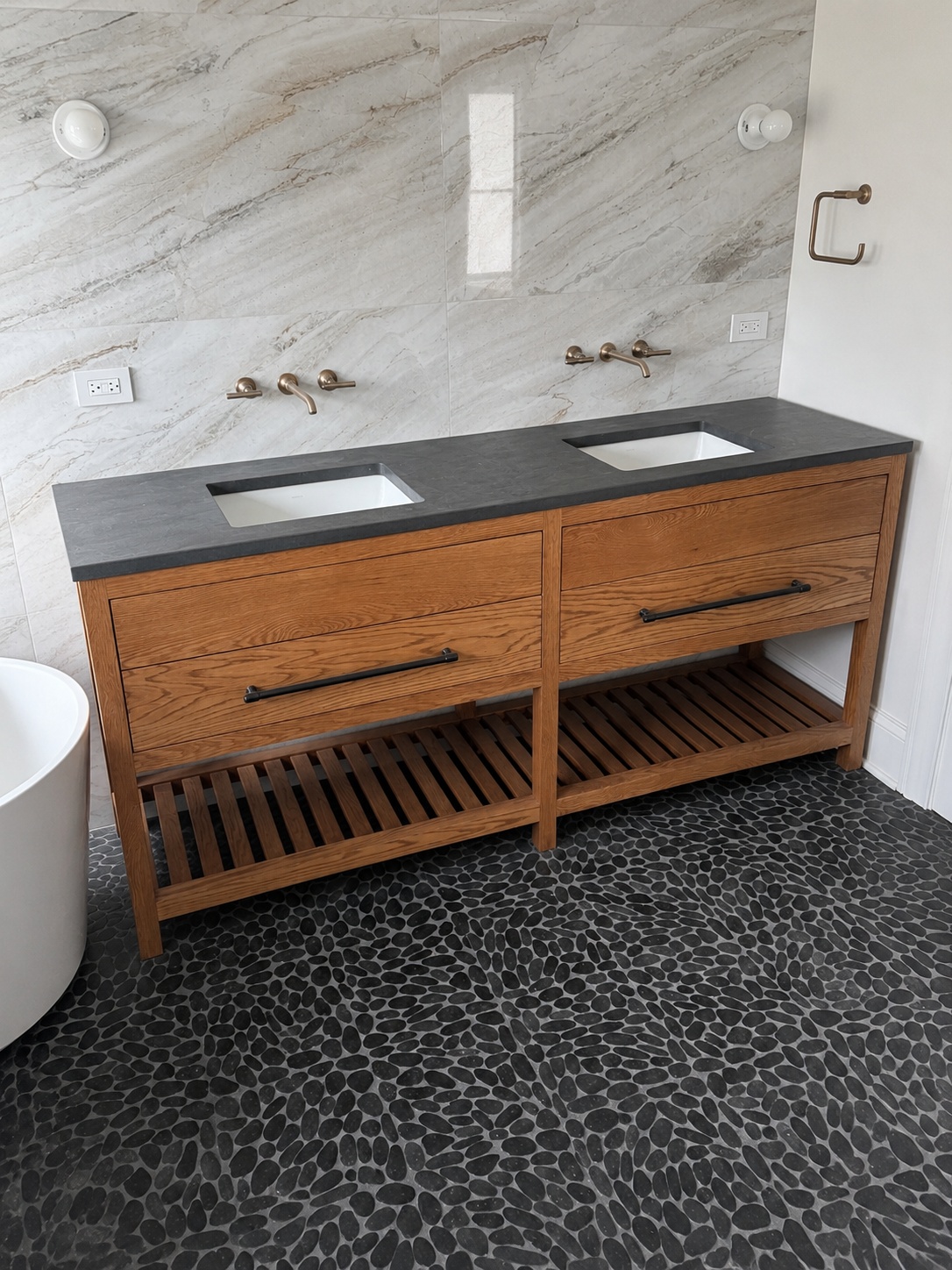 Natural oak double vanity with honed black stone top, brass wall faucets, marble tile wall, and pebble floor